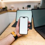 Mão segurando um smartphone em uma cozinha, ao lado de laptops sobre uma mesa de madeira.