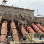 Usina hidrelétrica de grande porte com estruturas industriais e linhas de transmissão de energia.